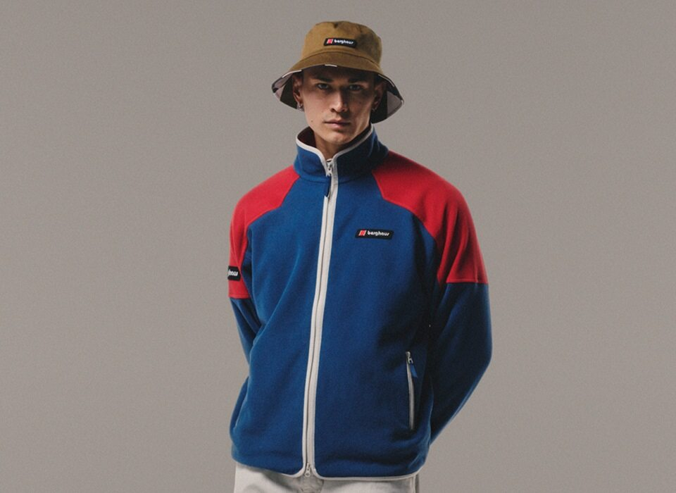 A man stood facing the camera with a wearing a khaki bucket hat on and a blue and red fleece facing towards the camera